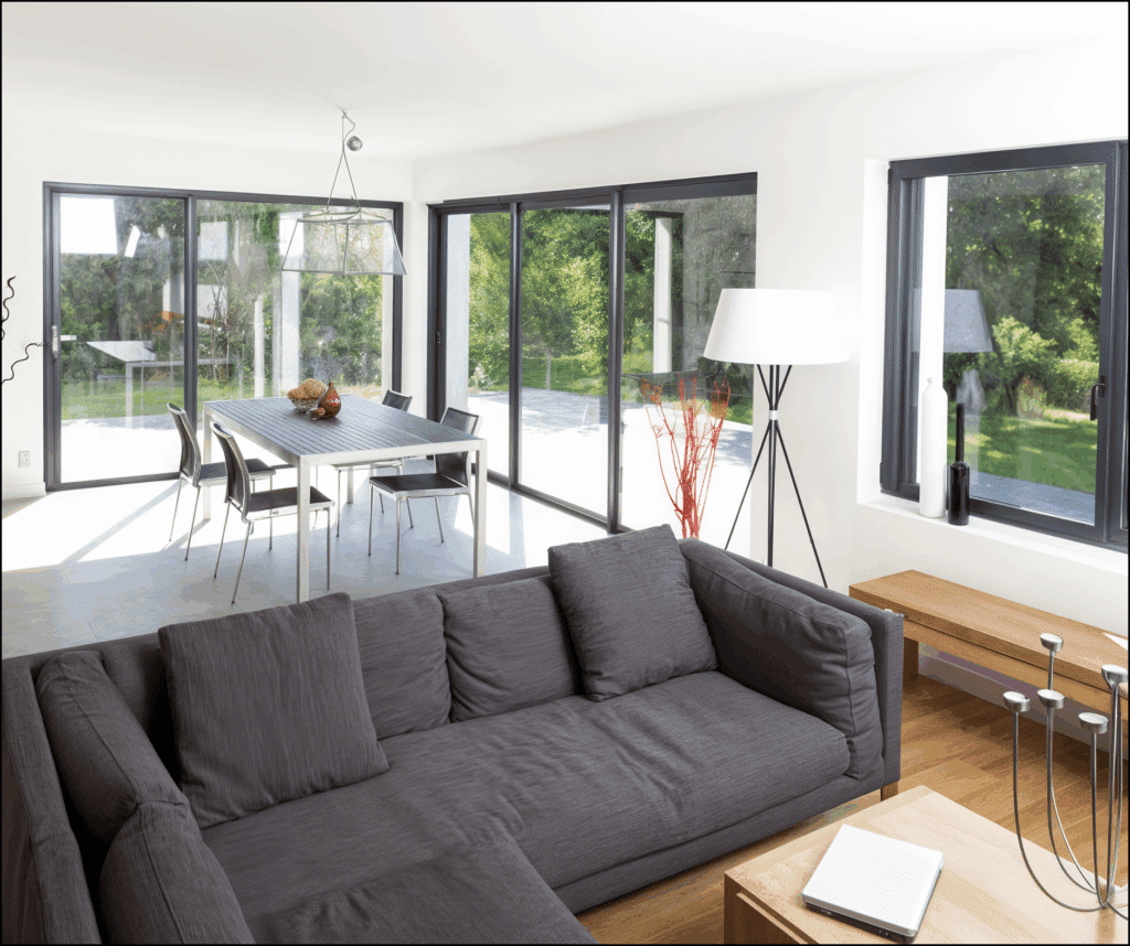 Salón comedor moderno con sofá gris y grandes ventanas a medida de Sabaya en aluminio oscuro que conectan el interior con el jardín